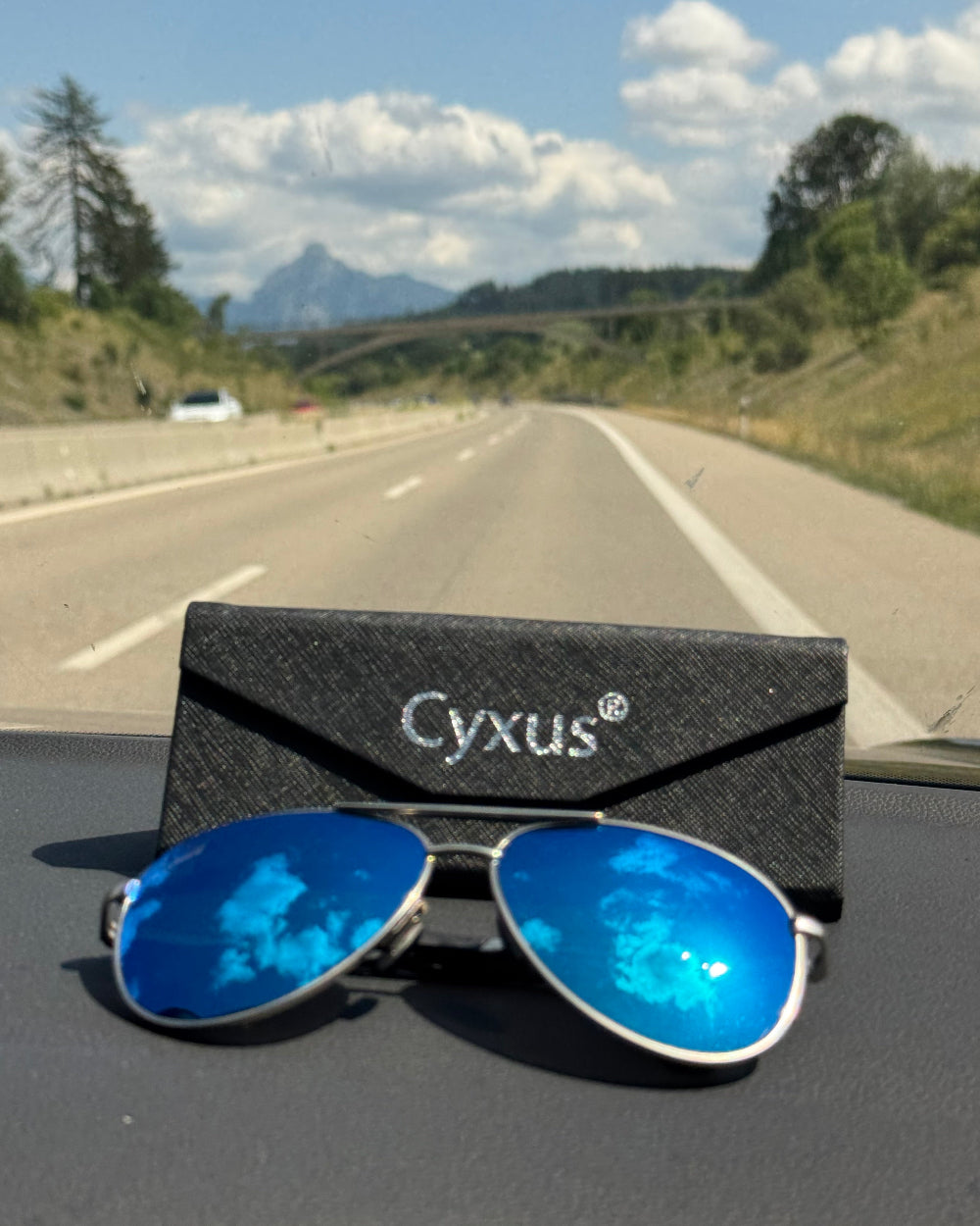 Blue sunglasses with a Cyxus case on a car dashboard, with a scenic road in the background.