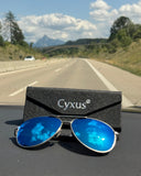 Blue sunglasses with a Cyxus case on a car dashboard, with a scenic road in the background.