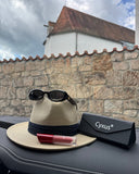 Sun hat, sunglasses, lipstick, and Cynxus case on a car dashboard with a stone wall and cloudy sky in the background.