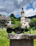 Sunglasses on a wooden post with a scenic background featuring a church and greenery.
